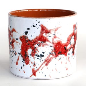Könnte beinhalten: Zylindrischer Keramik-Pflanztopf mit weißer Außenseite, verziert mit roten und schwarzen Spritzmustern. Der Innenrand ist warmbraun. Dieser dekorative Pflanztopf ist für den Innen- und Außenbereich geeignet.