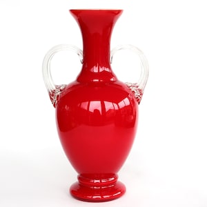 Op de afbeelding: Een rode glazen vaas met twee transparante glazen handvatten. De vaas heeft een rond lichaam en een uitlopende bovenkant.
