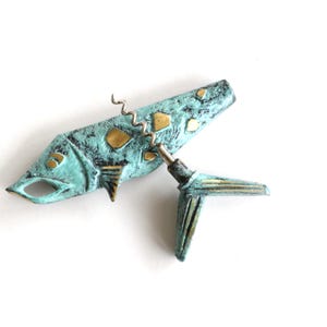 May include: A fish-shaped corkscrew with a textured, oxidized turquoise-green finish and polished gold-tone accents. The metal tool features a spiraled worm extending from the body, with a separate tail piece attached to the mechanism.