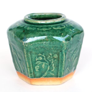 Op de afbeelding: Een zeshoekige keramische pot met een glanzend donkergroen glazuur en een zichtbare craquelé afwerking. De buitenkant is voorzien van reliëf bloemmotieven op elk zijpaneel. De basis is ongeglazuurd en toont een lichtbruine, matte kleitextuur.