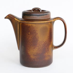 May include: A brown ceramic teapot with a lid and handle. The teapot has a spout for pouring and a rounded body. The color is a mix of brown and gold, with a speckled texture. The teapot is likely for brewing tea.