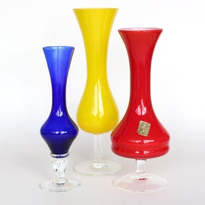 Op de afbeelding: Drie glazen vazen met uitlopende randen en heldere glazen stelen op ronde voeten. De linker vaas is kobaltblauw, de middelste vaas is felgeel en de rechter vaas is rood met een kleine rechthoekige sticker op het lichaam van het glas.