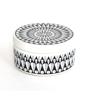 Op de afbeelding: Een ronde, witte metalen trommel met een zwart-wit geometrisch patroon. Het deksel heeft een stralend stervormig ontwerp, terwijl de zijkant van de container is versierd met een herhalend motief van gestileerde driehoeken en cirkelvormige figuren.