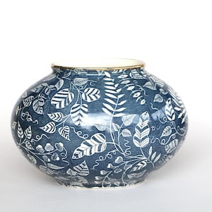 Op de afbeelding: Een ronde, blauwe keramische vaas met een wit bladerpatroon. De vaas heeft een gouden rand en een glanzende afwerking. De bladeren zijn gestileerd en gerangschikt in een herhalend patroon over het oppervlak.