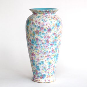Op de afbeelding: Een witte keramische vaas met een gespikkeld patroon in tinten roze, geel en blauw. De vaas heeft een lichtblauwe rand en een afgeronde basis.