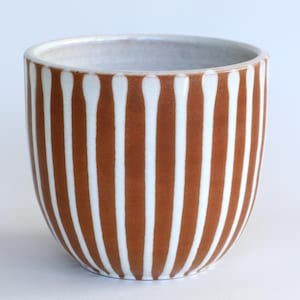 Op de afbeelding: Een keramische tumbler met een afgeronde vorm. De buitenkant heeft verticale strepen in wit en een warme bruine kleur. De binnenkant is wit. Deze handgemaakte beker is geschikt voor warme of koude dranken.