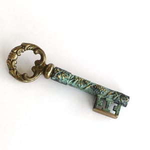 Könnte beinhalten: Ein dekorativer Metallschlüssel mit einem verzierten, messingfarbenen runden Griff und einem zylindrischen Schaft mit einer verwitterten, blaugrünen Patina. Der Schaft ist mit floralen Prägemustern versehen und der Bart hat ein rechteckiges Ausschnittdesign.