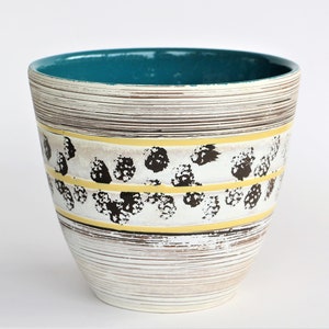 Op de afbeelding: Een keramische plantenbak met een turquoise interieur en een witte, bruine en gele buitenkant. De plantenbak heeft horizontale lijnen, een band met zwarte stippen en gele strepen. Het ontwerp is een mix van moderne en vintage stijlen.