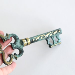 Könnte beinhalten: Ein dekorativer Metallschlüssel mit einer verwitterten, blaugrünen Patina und polierten goldfarbenen Akzenten. Der Schlüsselkopf hat ein verziertes, durchbrochenes Design, während Schaft und Bart komplexe, erhabene Rankenmuster zeigen.