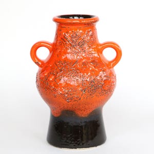 Op de afbeelding: Een keramische vaas met een getextureerde oranje body en twee lusvormige handvatten. De basis is zwart en de binnenkant is donker. De vaas heeft een mid-century modern esthetiek en staat op een witte achtergrond.