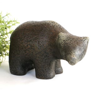 May include: A small, brown bear figurine with a textured surface. The bear is in a walking pose, with its head slightly lowered. The sculpture is made of a dark, earthy material, and is a decorative item.