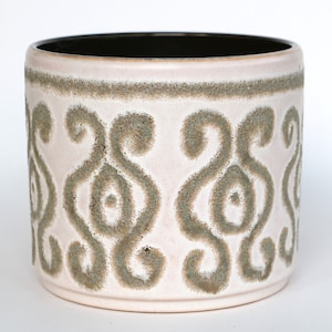 Op de afbeelding: Cilindrische keramische plantenbak met een lichtroze basis en een donkerbruin interieur. De buitenkant heeft een herhalend patroon van gestileerde, wervelende vormen in een gedempte groene kleur. Geschikt voor binnen of buiten.