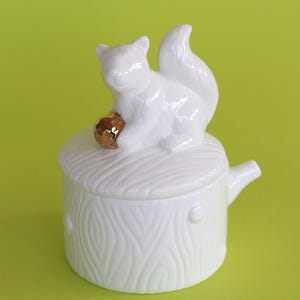 May include: A white ceramic lidded jar shaped like a tree stump with a squirrel figurine on top. The squirrel is holding a small, metallic gold-colored nut. The jar features a textured bark pattern on the exterior and a small spout on the side.