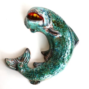 Op de afbeelding: Een keramieken vissculptuur in een gebogen C-vorm. Het oppervlak heeft een glanzend, gevlekt glazuur in blauwgroen en donkergroen met een textuur die op schubben lijkt. Het oog is opvallend en uitgevoerd in dieprood en geel.