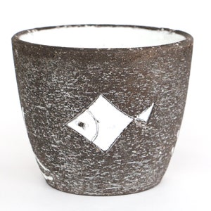 Puede incluir: Una taza de cerámica marrón oscuro texturizada con interior blanco. La taza presenta un diseño de pez blanco en forma de diamante en el exterior. La taza es adecuada para contener bebidas o como objeto decorativo.