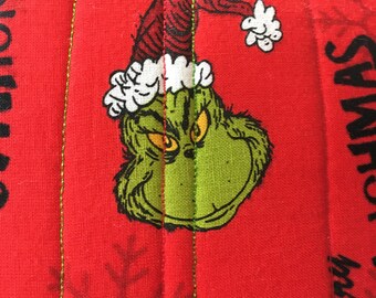 Peut inclure: Tissu rouge avec un personnage vert du Grinch portant un chapeau de Père Noël rouge et blanc. Le tissu a un motif matelassé et les mots "Joyeux Noël" et "Bonnes fêtes" sont imprimés dessus.