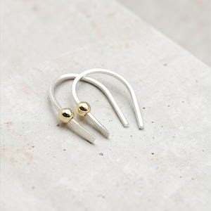 Puede incluir: Un par de pendientes de gancho plateados con detalles de bolas doradas. Los pendientes tienen un diseño sencillo y moderno, con un gancho curvado y una pequeña cuenta redonda dorada en la base. Los pendientes se muestran sobre una superficie gris claro.