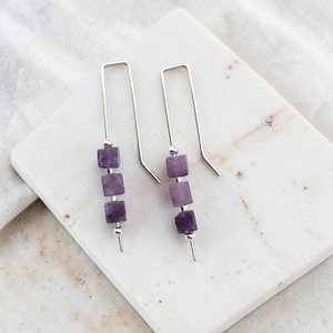 May include: A pair of silver-toned hook earrings, each featuring three small, square-shaped purple amethyst beads separated by tiny silver-toned metal spacers. The earrings have a long, straight wire design that extends vertically through the beads.