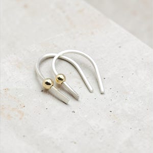 May include: A pair of U-shaped earrings crafted from silver-toned metal, each featuring a single small, polished gold-toned spherical bead positioned along one side of the curved wire structure, creating a simple and clean aesthetic design.