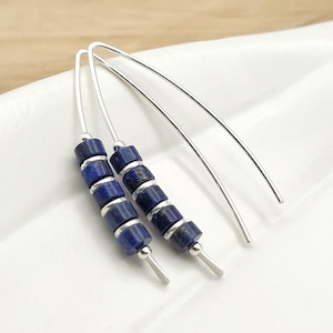 May include: A pair of silver-toned curved wire earrings, each featuring five cylindrical deep blue lapis lazuli beads separated by small silver-toned spacer beads, arranged in a vertical line along the lower curve of the wire.