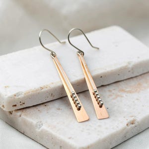 May include: A pair of dangle earrings featuring long, flat, rose gold-toned metal bars. Each bar is accented with a thin, vertical silver-toned wire adorned with four small, round silver-toned beads near the bottom. The earrings hang from silver-toned hooks.