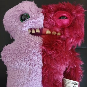 Op de afbeelding: Een knuffel in tweeën verdeeld, met de ene helft lavendel en de andere rood. De lavendelkleurige kant heeft een groot groen oog en witte tanden. De rode kant heeft een groot oog en tanden. Het speelgoed heeft een harige textuur.