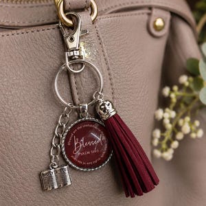 May include: A silver-toned keychain featuring a circular charm with a deep red background, the word Blessed, and the text Psalm 119:1. It includes a small silver book charm labeled Holy Bible and a long, deep red tassel attached to a metal clasp.