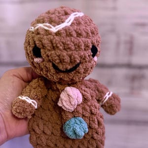 May include: A brown crocheted gingerbread man doll with white icing details and three colorful crocheted buttons. The doll has black eyes and a smiling mouth. The arms and legs are short and stubby.