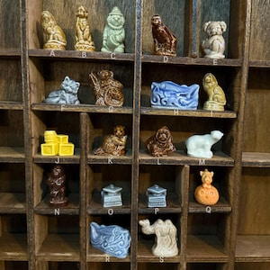 May include: A collection of small ceramic figurines displayed in a wooden organizer. The figurines depict various animals, including a lion, cat, bear, and monkey, in a range of colors. The organizer has multiple shelves, each holding a different figurine.