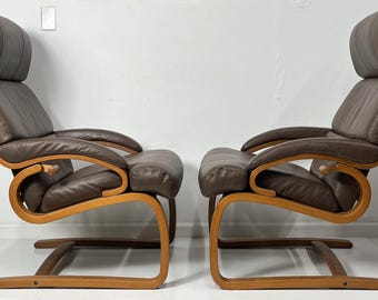 May include: Two high-back lounge chairs featuring dark brown leather upholstery and curved light-toned wooden frames. Each chair has a cantilevered base, integrated armrests, and a padded headrest, positioned side by side against a white wall.