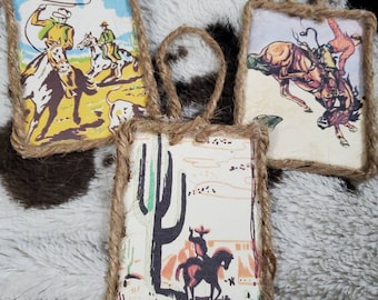 Puede incluir: Tres adornos rectangulares enmarcados con cuerda de yute gruesa. Uno muestra a un vaquero a caballo con un lazo, otro representa un caballo encabritado con un jinete, y el tercero presenta la silueta de una persona a caballo junto a un cactus grande.