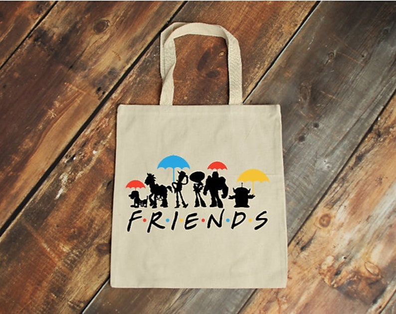 May include: A beige canvas tote bag with a graphic of cartoon character silhouettes under colourful umbrellas. The word "FRIENDS" is printed in black below. The bag has a handle for carrying.