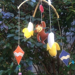 May include: A wooden baby mobile with a brown monkey, two yellow bananas, and two colorful diamond-shaped kites.