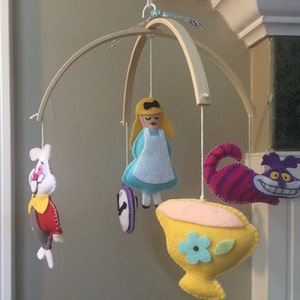 K&ouml;nnte beinhalten: Ein Holz-Babymobil mit Filzfiguren aus Alice im Wunderland. Das Mobile zeigt Alice, das wei&szlig;e Kaninchen, die Grinsekatze und eine Teetasse.