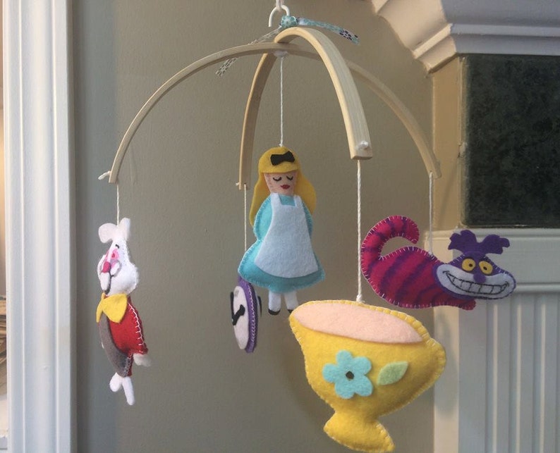 K&ouml;nnte beinhalten: Ein Holz-Babymobil mit Filzfiguren aus Alice im Wunderland. Das Mobile zeigt Alice, das wei&szlig;e Kaninchen, die Grinsekatze und eine Teetasse.