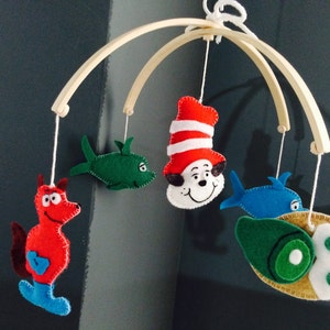 K&ouml;nnte beinhalten: Ein Holz-Babymobil mit Filztieren und Fischfiguren. Die Figuren umfassen einen roten Fisch, einen gr&uuml;nen Fisch, einen blauen Fisch und eine rote Katze mit Hut.