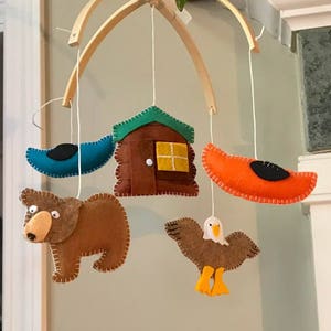 May include: A wooden baby mobile with felt animal and cabin shapes. The mobile features a brown bear, a brown eagle with yellow feet, a blue canoe, an orange canoe, and a brown cabin with a green roof and a yellow window.