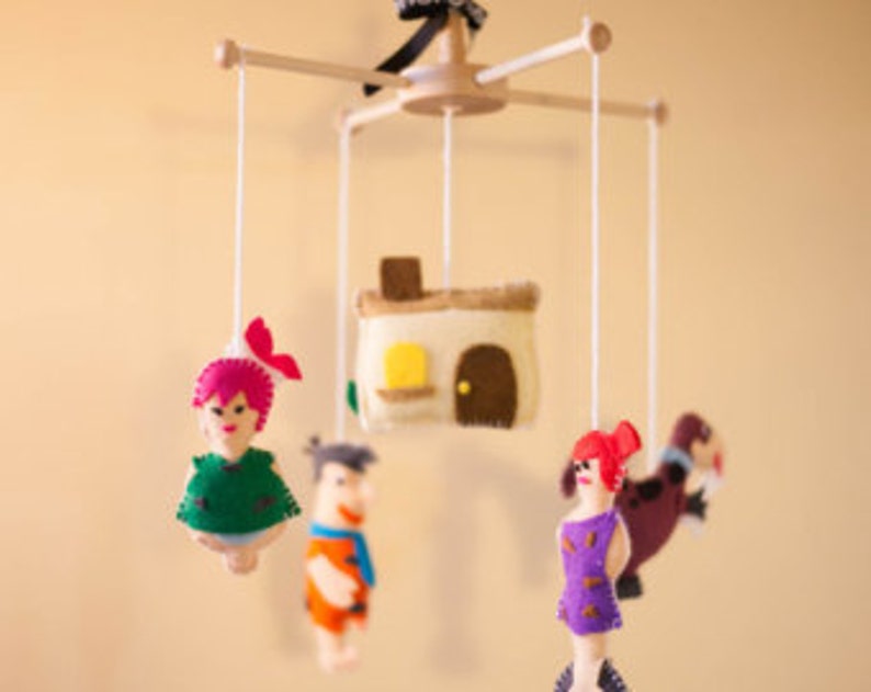 K&ouml;nnte beinhalten: Ein Holz-Babymobil mit Filzfiguren aus der Zeichentrickserie Familie Feuerstein. Das Mobile zeigt Fred, Wilma, Pebbles und ein Haus.