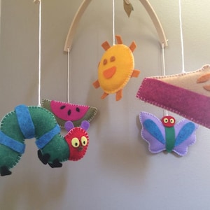 May include: A felt baby mobile with a green caterpillar, a red and green watermelon, a yellow sun, a purple butterfly with a green body and red eyes, and a pink and brown pie.