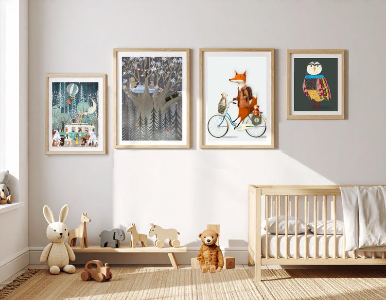 May include: Four framed wall art prints featuring whimsical animal illustrations, including a fox on a bicycle, a forest scene with a camper, a tree with animals, and a colorful owl, displayed above a wooden crib and a low shelf with wooden toys.