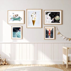 May include: A gallery wall arrangement of five framed art prints featuring space-themed illustrations. The prints depict a rocket ship with animals, a planet-topped ice cream cone, an astronaut painting a planet, a rocket on a moon, and a red rocket.