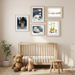 May include: A gallery wall arrangement of five framed art prints featuring whimsical illustrations of animals, a lighthouse, and a sailing ship, displayed above a light wood crib with a teddy bear and a plush giraffe toy sitting on the floor.