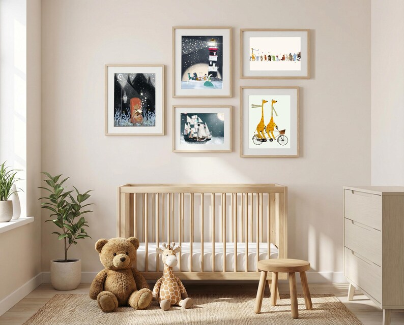 May include: A gallery wall arrangement of five framed art prints featuring whimsical illustrations of animals, a lighthouse, and a sailing ship, displayed above a light wood crib with a teddy bear and a plush giraffe toy sitting on the floor.