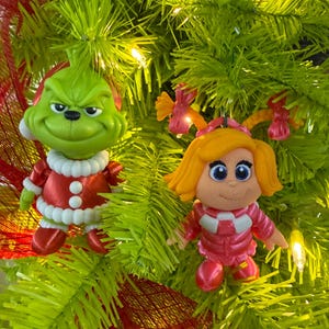 Könnte beinhalten: Zwei Christbaumschmuckstücke mit Figuren aus einem Weihnachtsfilm. Eines ist eine grüne Grinch-Figur in einem rot-weißen Weihnachtsmann-Anzug. Das andere ist ein Mädchen in einem rosa Outfit. Die Ornamente sind vor einem grünen Weihnachtsbaum angebracht.