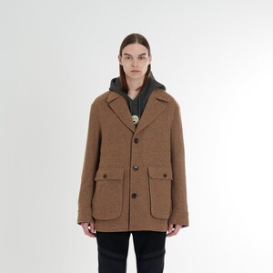 Op de afbeelding: Een bruin tweedjack met een knoopsluiting, twee klepzakken en een kraag. Het jack wordt gedragen over een donkergroen hoodie.