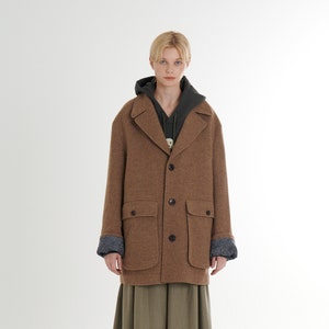 Op de afbeelding: Een bruine wollen jas met een kraag, knoopsluiting en twee grote zakken. De jas heeft een grijze en witte gebloemde voering aan de manchetten. Het model draagt een groene rok en witte laarzen.