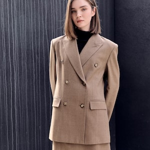 Op de afbeelding: Een beige double-breasted blazer en bijpassende rok. De blazer heeft vier gouden knopen en twee voorzakken. De rok heeft een wikkelontwerp. De persoon draagt een zwarte coltrui.