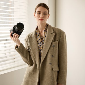 Op de afbeelding: Een beige double-breasted blazer met grote knopen en een textuurstof. De blazer wordt gedragen over een patroonjurk met een blauw en geel cirkelvormig ontwerp. Een zwarte camera wordt in de linkerhand gehouden.