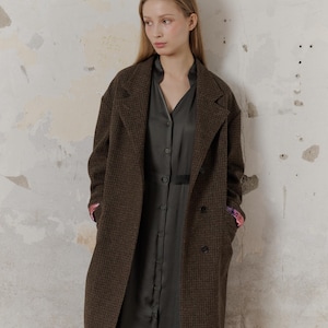 Op de afbeelding: Een bruin en zwart geruite jas met een dubbele rij knopen. De jas heeft een kraag en zakken. Het model draagt ​​een donkergroene jurk onder de jas.