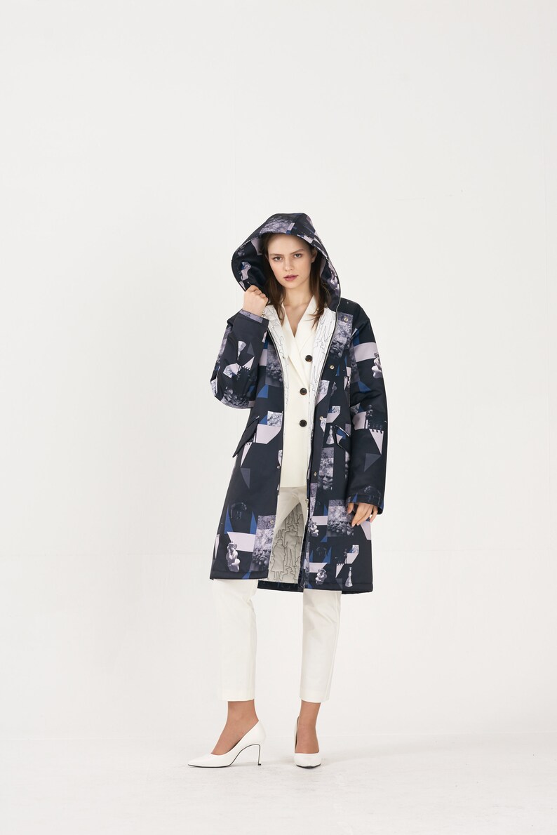 Puede incluir: Una chaqueta larga con capucha de color negro y blanco con estampado, con una cremallera blanca y un cuello blanco. La chaqueta se lleva sobre una camisa blanca abotonada y unos pantalones blancos.